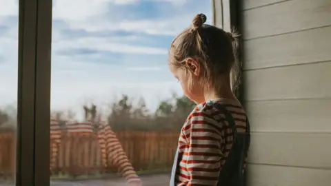 Getty Images Little girl with bunches looking out a large window, inside a green wooden hut. 