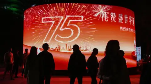 Getty Images People walk past a giant screen outside a shopping mall which displays a sign marking the 75th anniversary of the founding of the People's Republic of China, on the third day of a week-long National Day holidays in Beijing on 3 October, 2024.