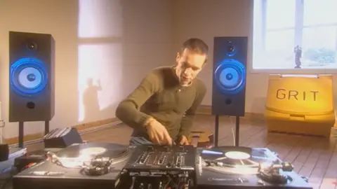 Martyn Bennett sits at a vinyl mixing deck, flanked by large speakers on stand  in a room with a large yellow street grit bin in below the window. a statuette is casting a shadow of a waving figure on the wall.