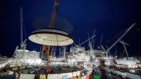 PA Media A giant crane lifts the huge dome roof onto the reactor building at Hinkley Point C. It's dark but the forest of cranes is lit up.