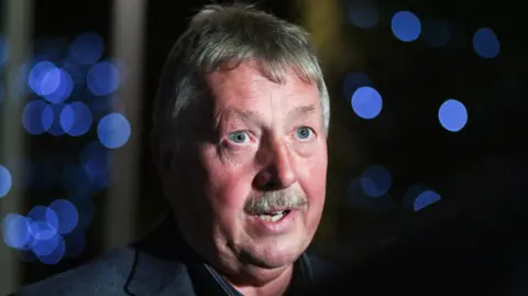 Reuters Headshot of Sammy Wilson speaking, looking slightly off camera. In the background some lights that are out of focus.