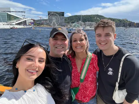 Jeannie Ambrose Jeannie Ambrose, her partner and two children are stood in front of a marina. They are all smiling at the camera. In the background there are boats and a Ferris wheel