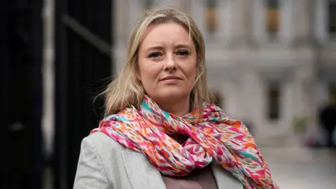 PA Media Máiría Cahill stands in front of the camera. She has blonde hair, a grey jacket and a colourful scarf. Her expression is neutral.