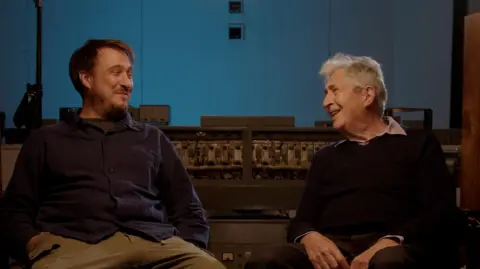 Reverb Hamish and Malcolm sit beside each other smiling at one another. They are sitting in a music studio with sound equipment behind them as well as a recording booth. Hamish has dark short hair, is wearing a blue shirt and brown trousers. Malcolm has grey short hair and is wearing a black jumper with a light shirt on underneath.