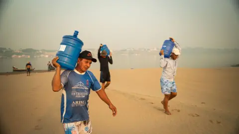 A group walk from the river across sand with water bottles 