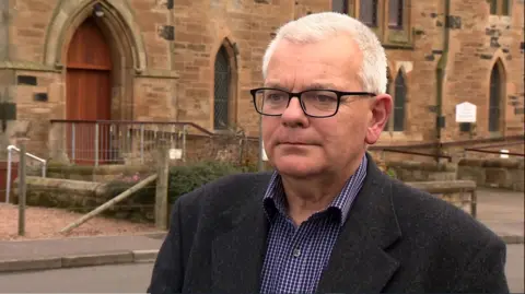 A man with grey hair wearing glasses, a dark suit and a blue chequered shirt standing in front of a church