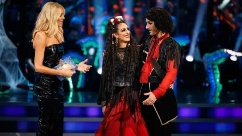 BBC/PA A woman in a black strapless dress smiling as she addresses a woman in a red and black dress and her partner. The woman in red has a skull and flower headpiece and a long black wig. Her partner is wearing a black jacket with gold trim and red accents. The couple is standing on a stage with a blurred background of lights and a crowd.