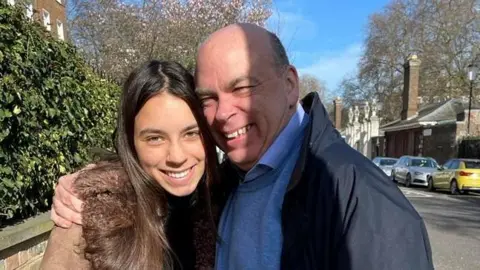 Reuters A photo of Hannah Lynch with her father Mike Lynch. Mr Lynch holds his daughter in an embrace as they smile at the camera. A road can be seen behind them with cars parked nearby and several buildings. Miss Lynch has long brown hair and is wearing a brown coat with a furred hood. Mr Lynch is wearing a navy coat with a blue jumper and shirt underneath.