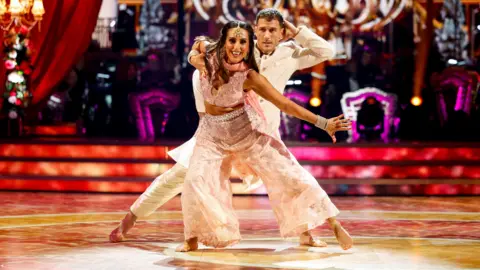 BBC/PA Dr Punam Krishan mid dance on the Strictly dancefloor, she is wearing a pink jewelled traditional Bollywood costume. She has her left arm and right leg extended. Her dancing partner Gorka Marquez stands behind her wearing a white Bollywood outfit.