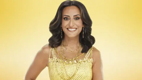 PA Media This image shows Strictly Come Dancing contestant Dr Punam Krishan wearing a yellow sequined dress and lots of gold jewellery. She has brown hair and gold eye makeup and is smiling at the camera. The photo has a solid yellow/gold background.