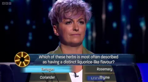 Woman with pink hair and a brown jacket looking serious as she ponders a quiz question on a game show.