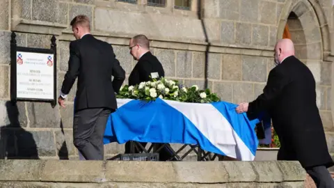 PA Media Three men dressed in black carry the coffin, topped with flowers, inside a church 