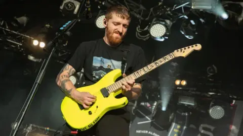 Getty Images A man with short hair and a beard wears a look of concentration as he plays notes on an electric guitar with a neon yellow body. He's got heavily tattooed arms and the fretboard of his guitar has 