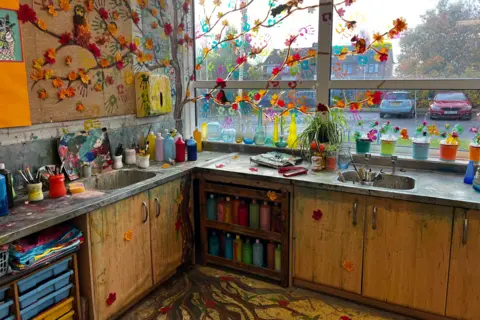 Josh Parry / BBC The corner of an art classroom used for filming Heartstopper. The counter tops and sinks are covered in bottles of paint and there is a mural of a tree with autumn leaves painted on the wall and along the window, its roots painted on to the floor