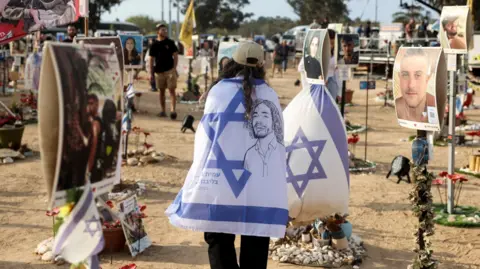 Reuters A woman walks while wearing a cream baseball cap and draped in an Israeli flag, as people visit the site of the Nova festival