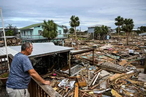 CHANDAN KHANNA/AFP A man inspects the damage of his home after Hurricane Helene. The image shows debris from houses as far as the eye can see.