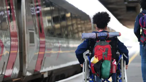 Getty Images A woman in a wheelchair moves along a train platform with luggage in tow