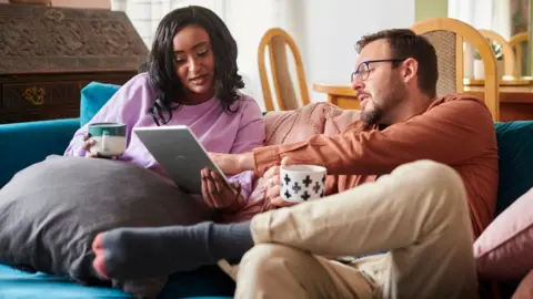 Getty Images A man and a woman sit on a sofa looking at a tablet computer. Both have mugs in their hands and are looking intensely at the screen.