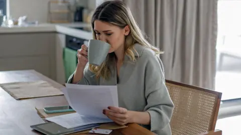 Getty Images A woman sits in a chair at home looking at paperwork and drinking a cup of tea