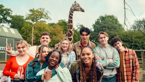 Netflix/Heartstopper Season 3 The cast of Hearstopper smile at the camera while filming on location at a zoo. A giraffe and trees are visible in the background.