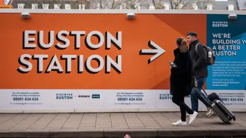 PA Media Two people walk with suitcases walk past construction hoarding at the HS2 construction site. The hoarding has information on it, including Euston Station written in large print with an arrow pointing commuters in its direction.