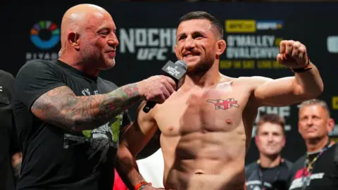 Getty Images Shirtless MMA fighter Merab Dvalishvili is interviewed by Joe Rogan, wearing a black T-shirt and holding a microphone, at UFC 306 in Las Vegas, Nevada in September