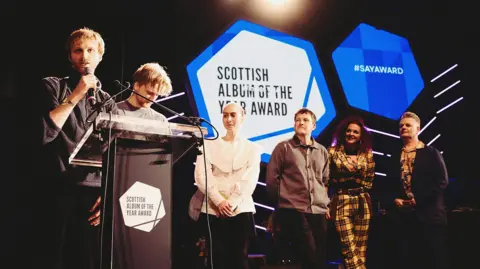 SAY Award Three members of redolent, two male and one female, stand at a podium in front of a large awards logo with other guests at the side of the stage