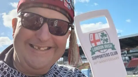 Nige Eaton Nige Eaton in a fez and sunglasses, posing next to a London Irish Supporters Club 20th Anniversary cut-out sign