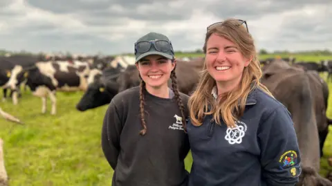 BBC Emily and Georgie Paul stand in front of their cattle, on a big grassy field. They are both smiling, and have sun on their faces