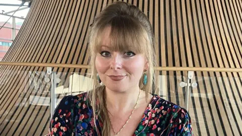 Delyth Jewell has long blonde-brown hair and is wearing a patterned dress. She is sitting in the public area of the Senedd, which surrounds the wooden funnel-shape of the Senedd chamber's ceiling, pictured behind her.