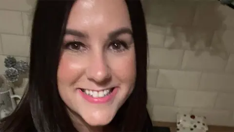 X Lucy Connolly, who has dark, shoulder length hair is smiling at the camera. She is standing in front of a white tiled wall indoors.