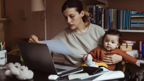 Getty Images Female professional wearing a grey jumper balances her baby while sitting down at a desk at home where she is reading a document