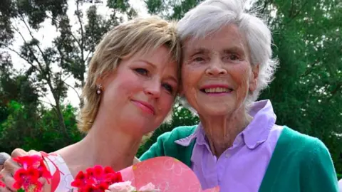 Sally Magnusson  Sally Magnusson (left), holding red and pink flowers, with her mother Mamie who is wearing a purple blouse and green cardigan. They are looking in the same direction and their heads are touching. There are green trees behind them. 