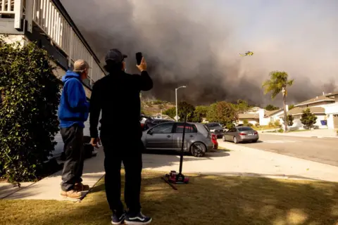 Getty Images Two men, one wearing a blue hoodie and beige cap and another wearing a black top and black cap holding up a camera phone, watch a firefighting helicopter fly above a plume of smoke