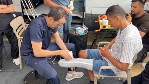 Rizeq Tafish sits on a chair whilst a Jordanian doctor wraps the stump of his leg in a white bandage, whilst a few people are stood or sat around them. 