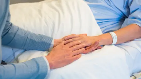 Getty Images A man holds the hand of a woman lying in a hospital bed