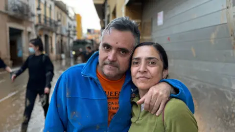 Juan Jose puts his arm around his sister, Lourdes Montane, standing in a muddy street with a woman in the background wearing a mask