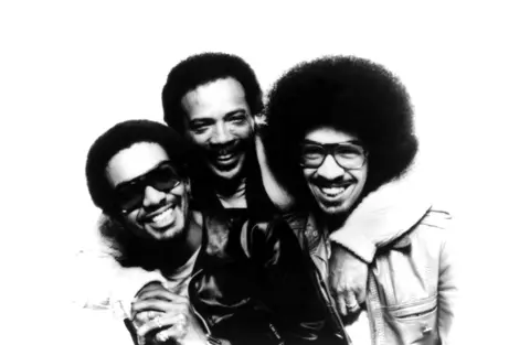 Getty Images Louis Johnson, Quincy Jones and George Johnson smile at the camera as they embrace