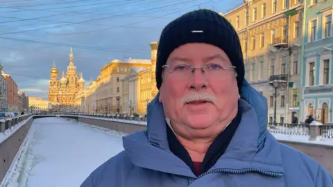 Facebook Headshot of David Crisp taken in St Petersburg. He has a grey moustache and rimless glasses and is wearing a black woollen beanie hat, a blue puffer coat, with a black jumper and other layers underneath. He is standing on a bridge over a frozen canal with an ornate church in the distance and buildings lining the canal. The sky is blue with soft sunlight on some of the buildings. 