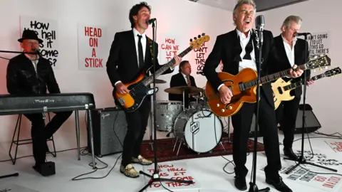 Danny Clifford Glen Matlock, playing a guitar and singing, performing with the four other members of his band, who are on keyboards, bass, guitar and drums. All are dressed in black and white, and there are slogans written on the wall and on the floor, such as 'head on a stick'