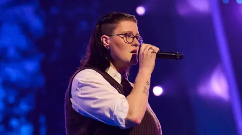 Shutterstock Hollie Peabody performing on The Voice. Hollie has long dark hair and an undercut above their ears. They wear a maroon knitted vest over a white shirt, their sleeves rolled up to their elbow revealing two tattoos: a small star and crossed plasters. They have a serious expression as they hold the mic to their face. 