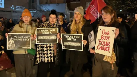 BBC A group of women stand on a winter's evening in Dublin, as part of a rally in support of Nikita hand who won a civil case against Irish mixed martial arts (MMA) fighter Conor McGregor. They are wearing signs saying 'thank you nikita'.