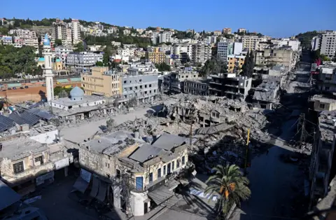 Reuters The destroyed market in Nabatieh. Grey debris from a pulverised structure covers a central square, which is lined by buildings with their windows blown out. High-rise buildings stand on the hills in the background. 