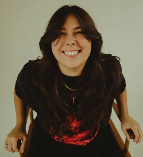 Nae Gigi Perez smiles while looking at camera while sitting in a chair against a neutral background, in a promotional image for her music.