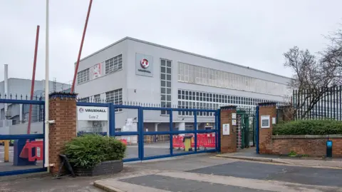 Getty Images The entrance to the assembly plant of the automotive manufacturing company IBC Vehicles Ltd, operating under Vauxhall Luton and producing light commercial vehicles, in December 2021. It shows blue metal gates, red barriers, and a grey building with lots of windows 