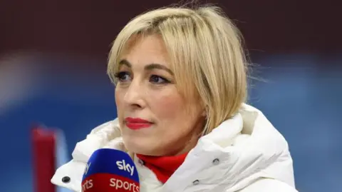 Getty Images Kelly Cates presenting on Sky Sports. She has blonde hair, and is wearing a white puffa jacket