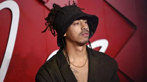 Getty Images Jordan Stephens wearing a hat with parts of his hair on show. He is wearing a baggy suit jacket without a top underneath, as well as a necklace. 