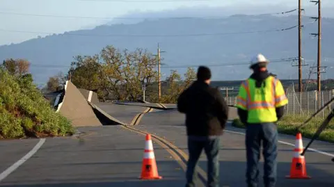 Getty Images A Pacific Gas and Electric worker looks at a road that buckled when it was undermined by a mudslide during a Bomb Cyclone storm in the week on March 23, 2023 in Novato, California