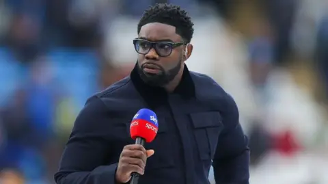 Getty Images Micah Richards presenting for Sky Sports, he has short black hair and is wearing a utility style navy coat and black glasses