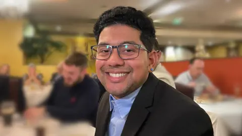 Ben Schofield/BBC Deeshen Ruttun looking direct to camera and smiling. He has black hair and dark eyes and is wearing black-framed glasses, a black jacket and a blue shirt. Other diners in a restaurant can be seen out of focus behind him.